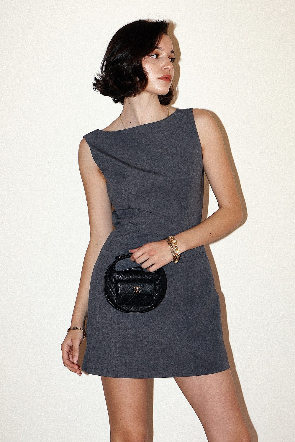 Model wearing a charcoal gray draped sleeveless mini dress with an asymmetrical bodice and mini-length hemline, styled with a black handbag.