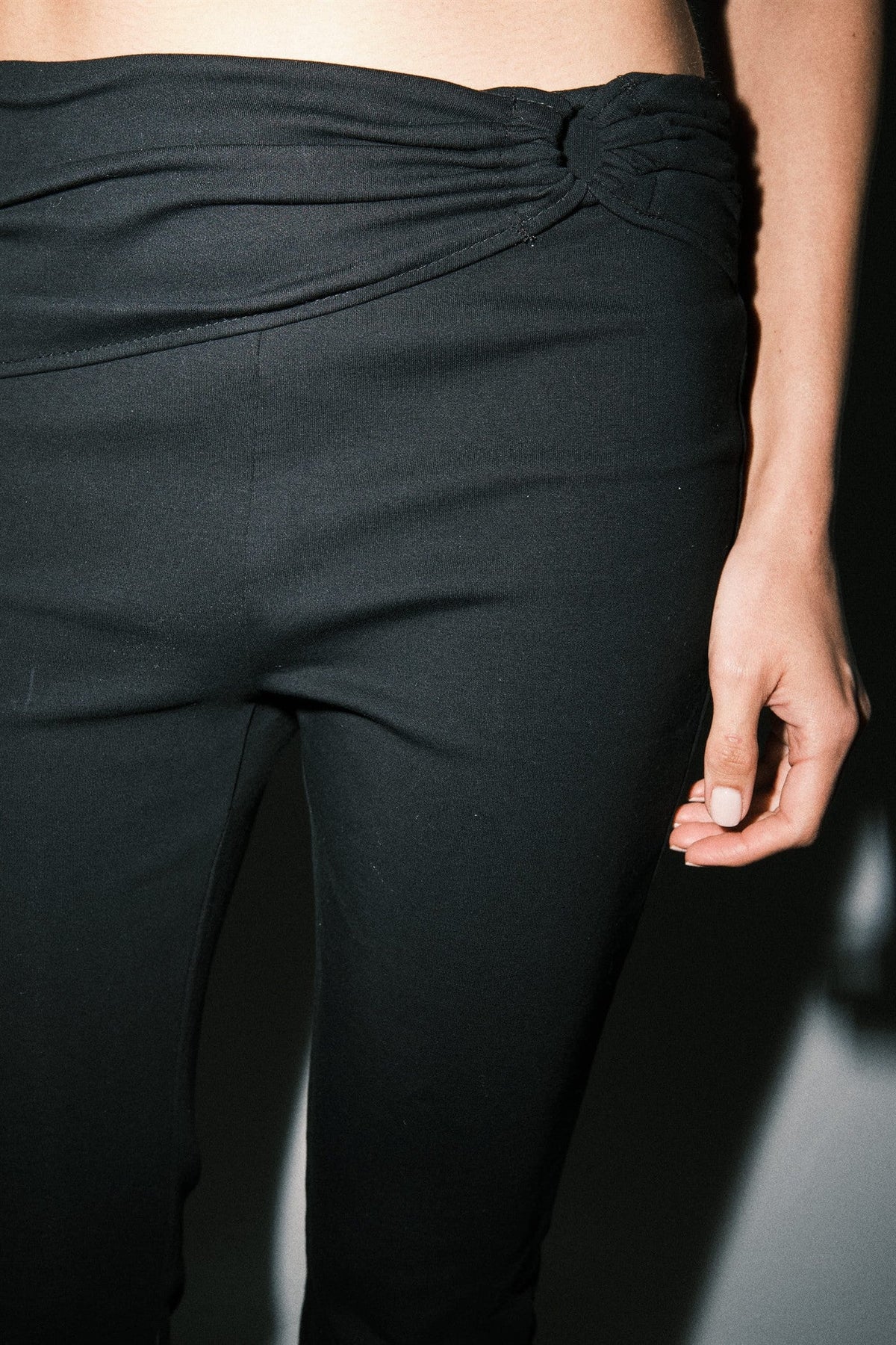 Close-up of black capri pants highlighting the asymmetrical draped waistband and gathered knot detail at the hip.